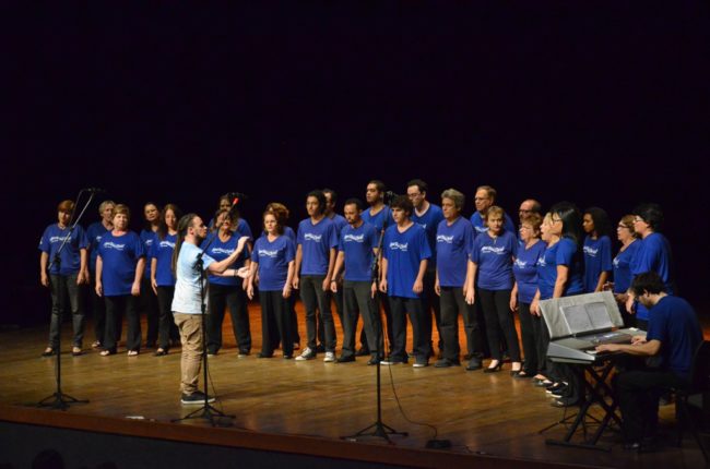 #PraCegoVer - Coral em cima do palco sendo regido por maestro